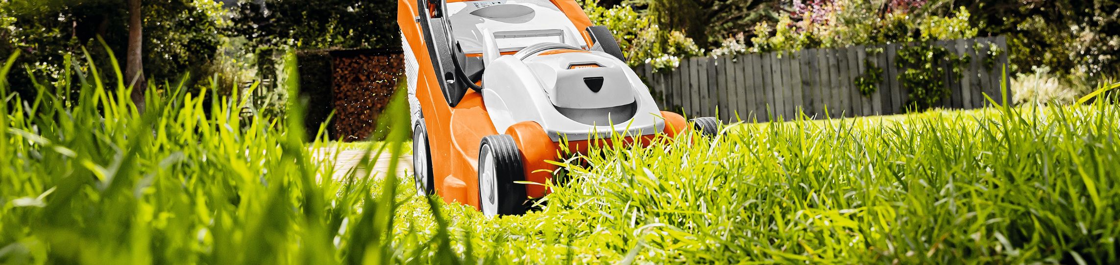 erson mäht mit einem STIHL Akku-Rasenmäher über eine grüne Rasenfläche, im Hintergrund ist ein Holzzaun, sowie grüne Büsche zu sehen.