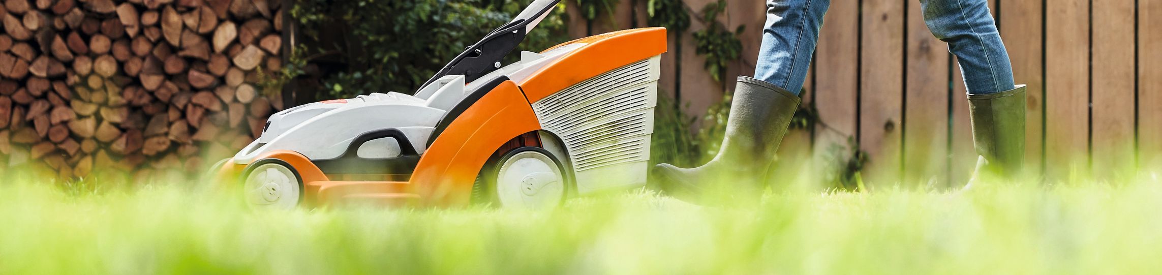 Person mäht mit einem STIHL Akku-Rasenmäher über eine grüne Rasenfläche, im Hintergrund gestapeltes Kaminholz und ein Holzzaun.