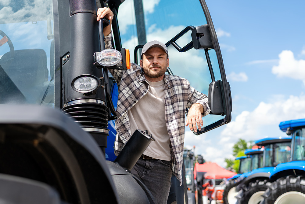 Ein Mitarbeiter sitzt auf einer New Holland Landmaschine im Vordergrund. Im Hintergrund stehen mehrere weitere Traktoren aufgereiht auf dem Betriebsgelände.