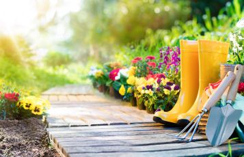 Gelbe Gummistiefel und Gartengeräte vor blühendem Beet zur Gartenpflege.