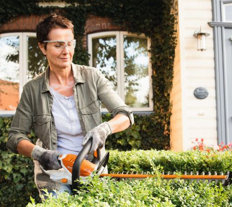 Eine Frau schneidet mit einer STIHL Heckkenschere eine Hecke und tragt dabei eine Schutzbrille und Arbeitshandschuhe.